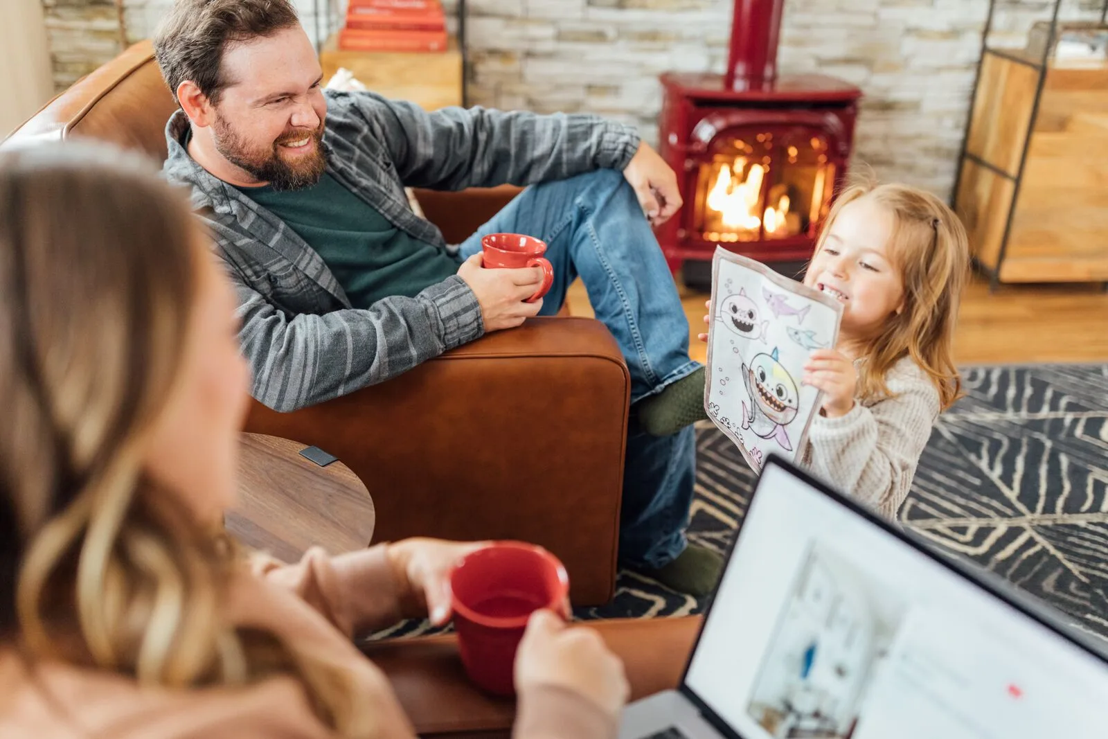 A dad with a mug and his daughter with a picture book beside a red Vermont Castings Stardance gas stove (manufacturer lifestyle photo)