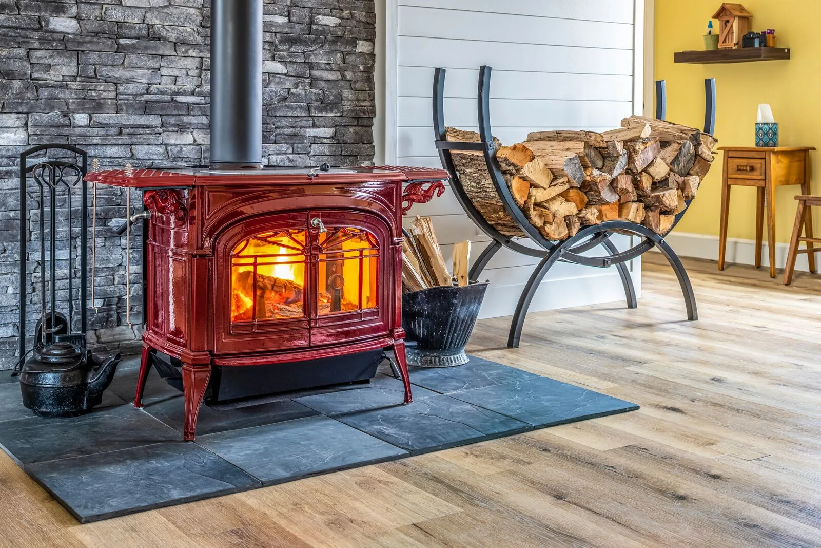 A Bordeaux-red Vermont Castings Encore wood stove on a slate hearth beside a log rack (manufacturer lifestyle photo)
