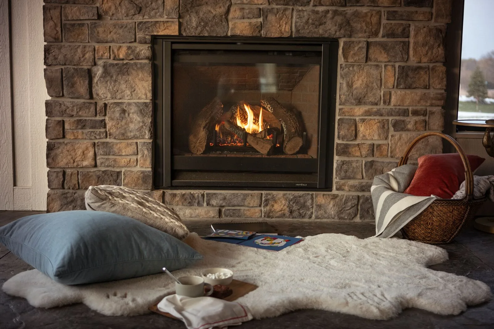 A gas fireplace burning in a stacked-stone wall beside a window, a tray of mugs on the ottoman (manufacturer lifestyle photo)